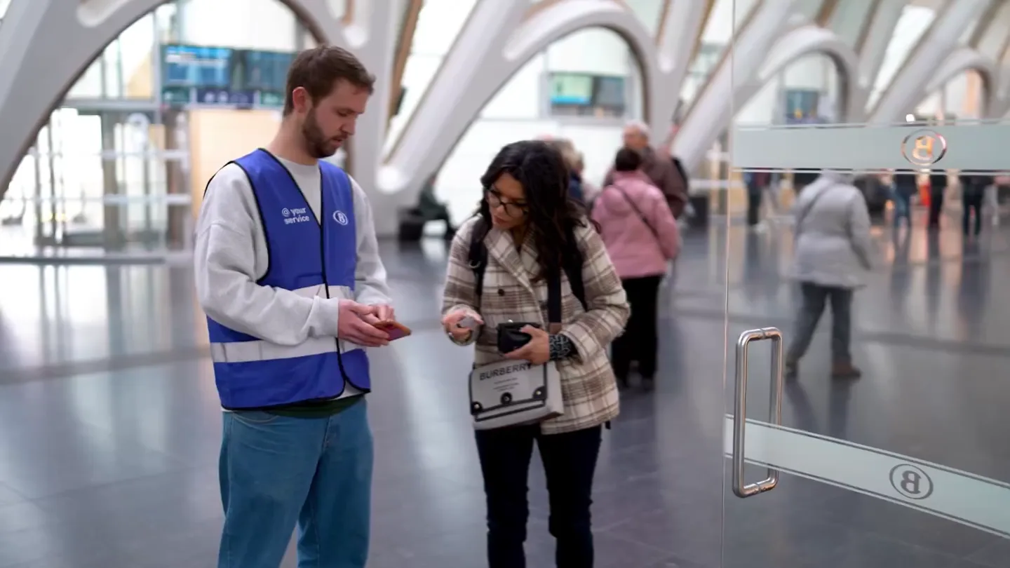 Host in blauw 'at your service'-hesje helpt een reizigster in een NMBS-station